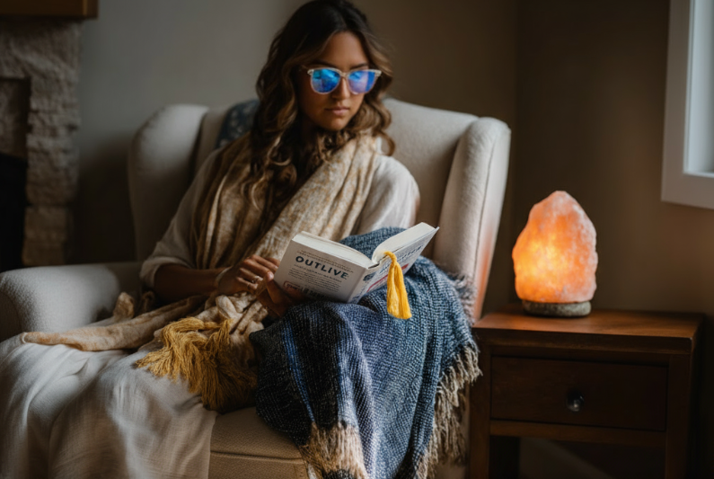 A woman with cool blue light glasses reads outline by peter attia at home next to a Himalayan salt lamp